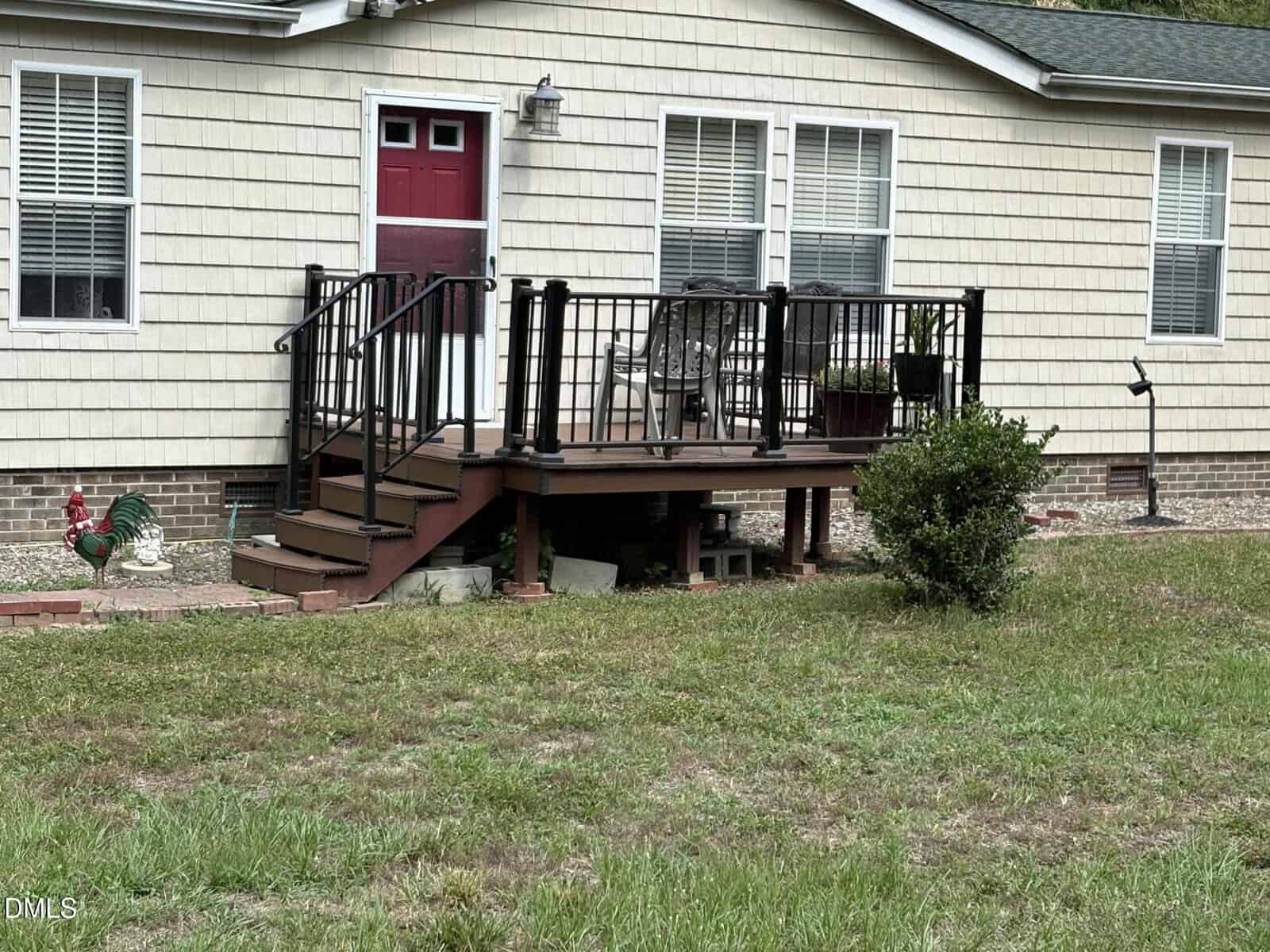 A small porch with black railings and wooden steps leads to a red front door of a beige house. Patio chairs and potted plants sit on the porch, and a bush and rooster statue are in the yard below.