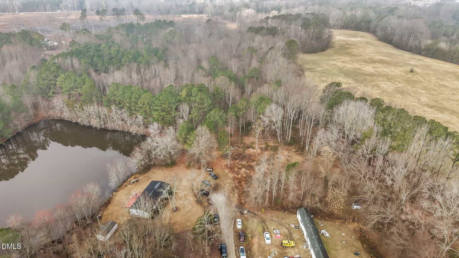 Aerial view of a rural property with a pond, a house, scattered vehicles, trees, and an open grassy field, surrounded by dense woods under an overcast sky.