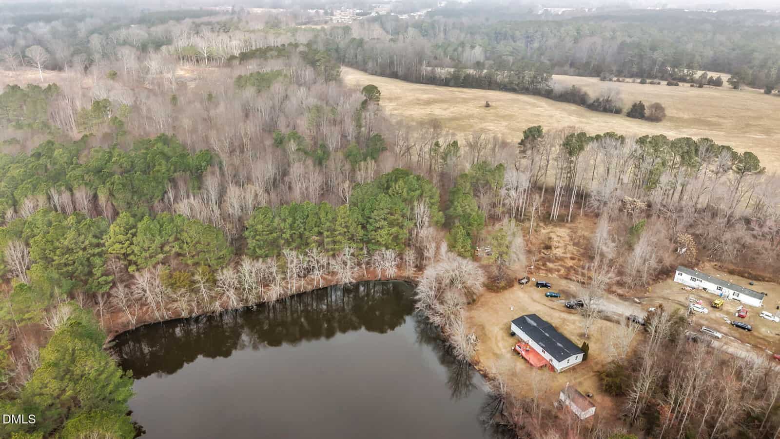 Aerial view of a wooded area with a pond in the foreground, a long building with red trim near the pond, and open fields and more trees in the background under a cloudy sky. Several vehicles are parked near the buildings.