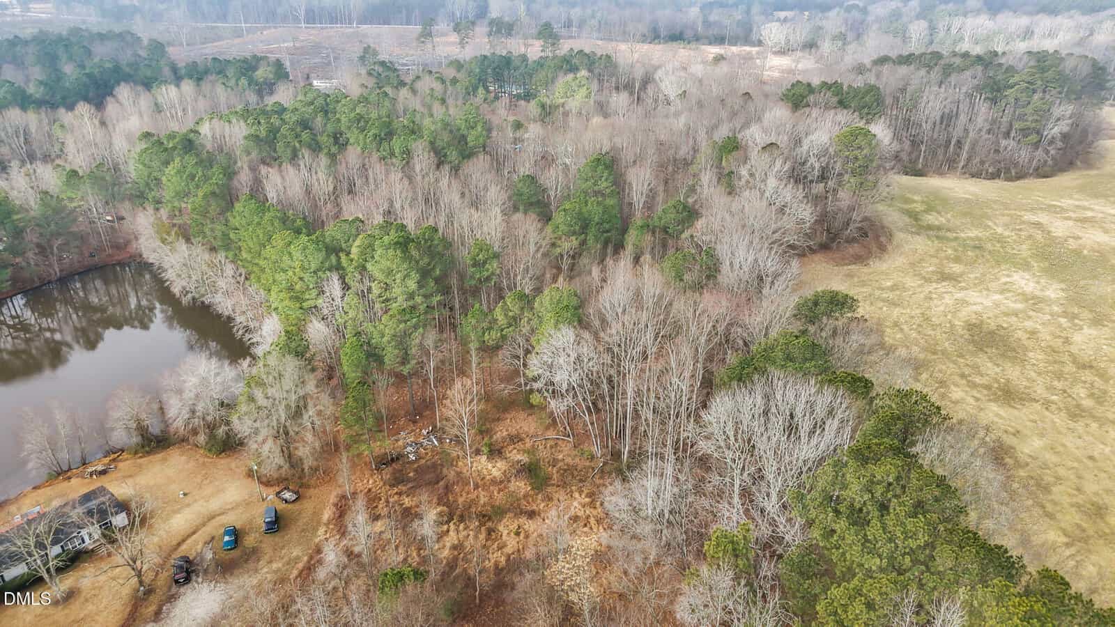 Aerial view of a forested area in winter with a mix of green pine and leafless trees, a small pond, scattered vehicles, open grassy fields, and distant roads visible in the background.