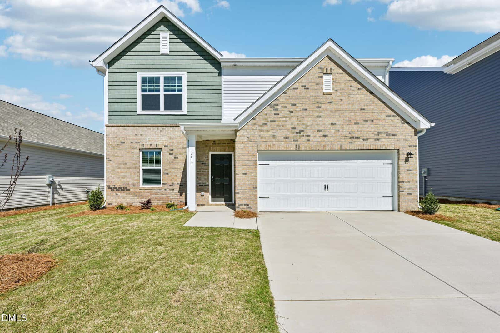 A two-story house at 216 Tar Banks Drive in Louisburg features green and beige brick siding, white trim, a double garage, and a concrete driveway. The neatly trimmed lawn has minimal landscaping under blue skies with scattered clouds.