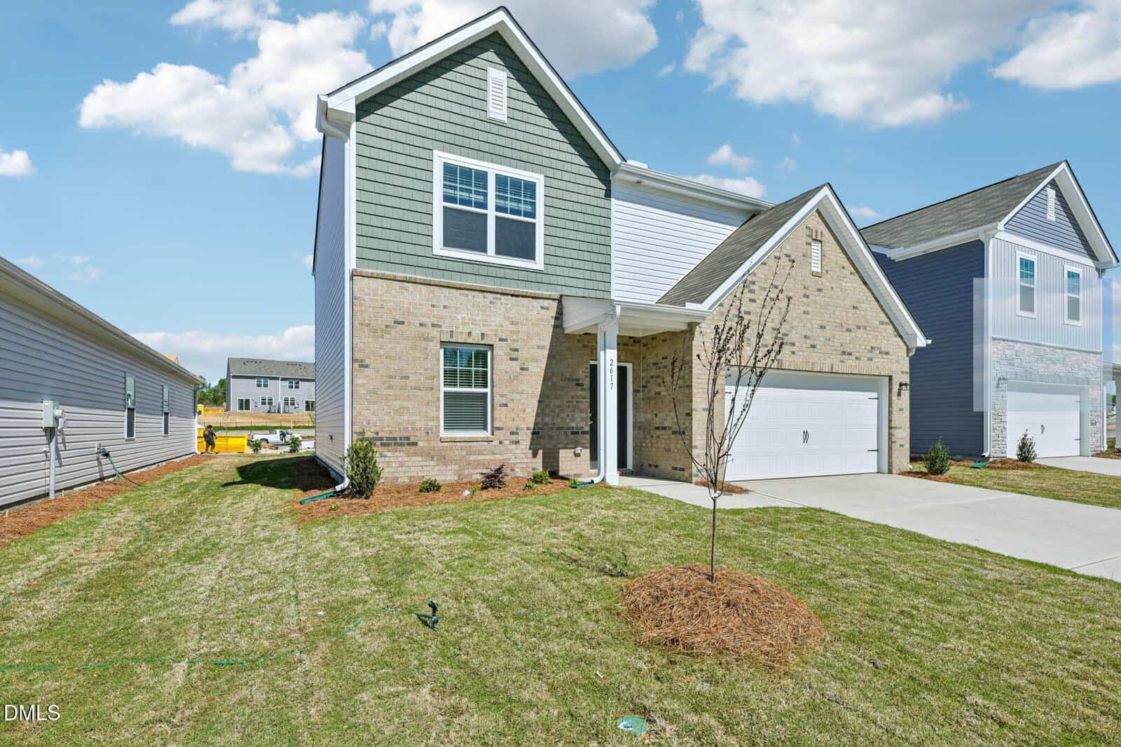 A modern two-story suburban house with brick and green siding, white trim, and a double garage. The front yard has fresh grass, small shrubs, and a young tree. The sky is blue with scattered clouds.