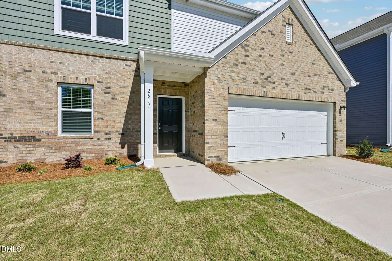 A modern two-story house with a brick exterior, a black front door, white trim, double garage doors, and neatly landscaped lawn and shrubs. The house number 2617 is visible near the entryway.