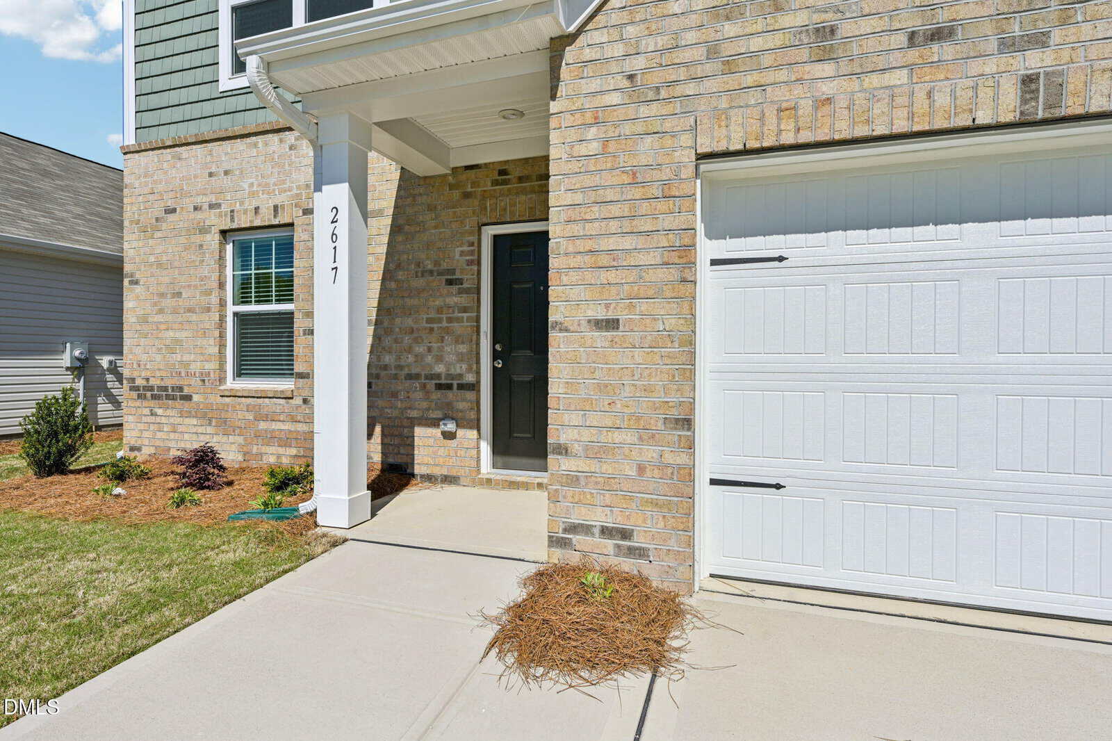 Front entrance of a modern brick house with a white pillar displaying the number 2617, a black front door, attached garage, and small landscaped area with shrubs and mulch.