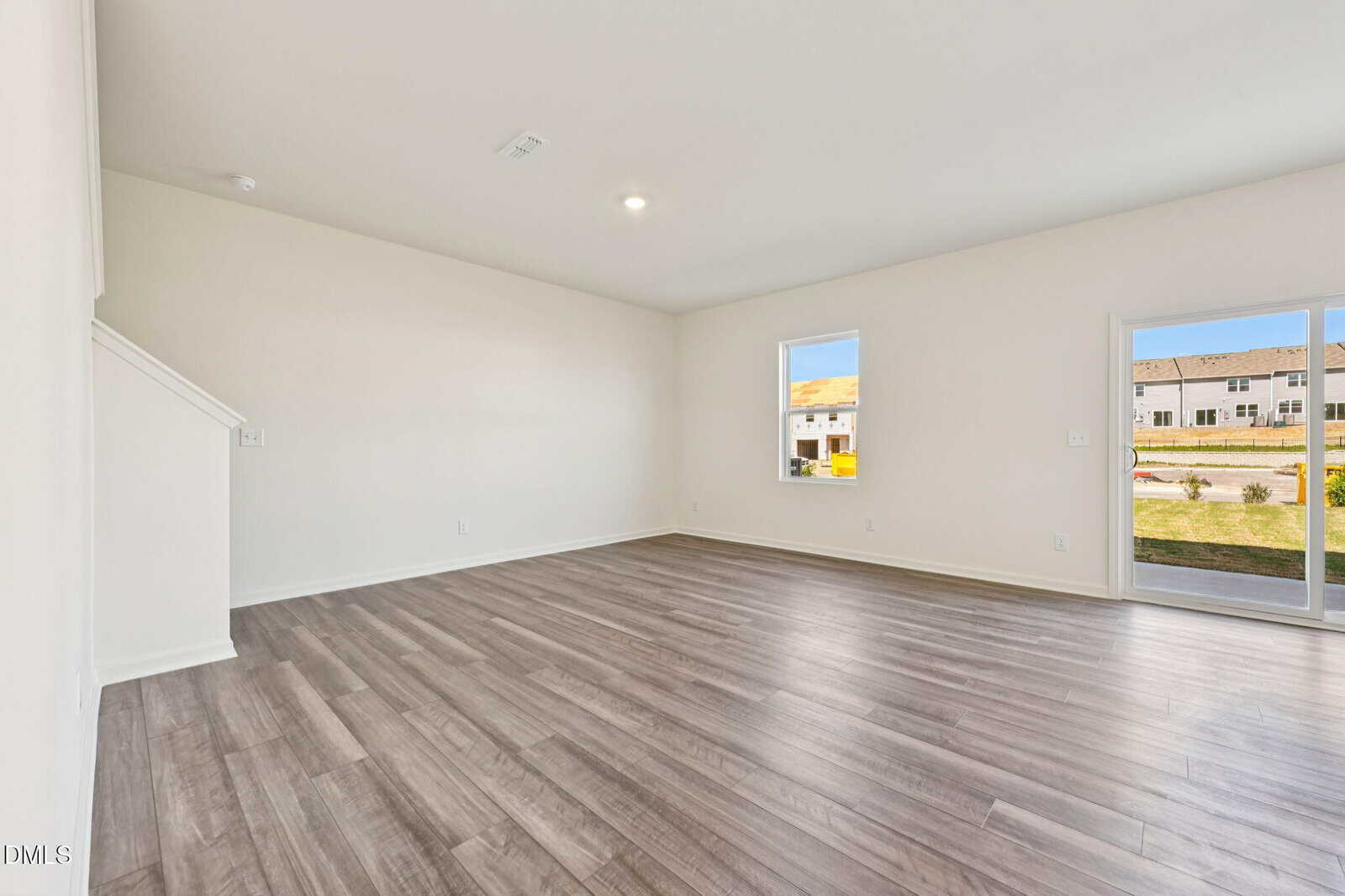 Empty living room with light gray wood flooring, white walls, and ceiling. Room has two windows and a glass door; outdoor area and other buildings visible through them. Staircase is partially visible on the left.