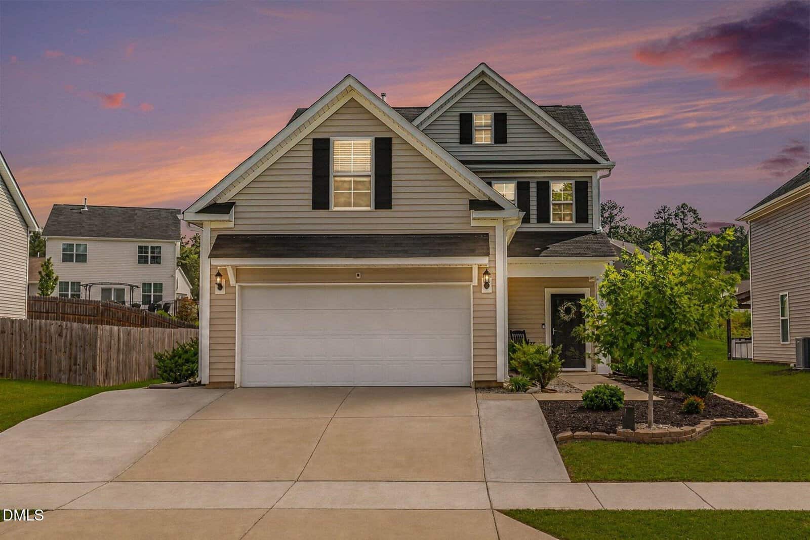 A two-story suburban house at 30 Kimberling Drive in Franklinton features beige siding, black shutters, and a double garage. Set against a colorful sunset sky, the front yard boasts a small tree, shrubs, and well-kept landscaping.