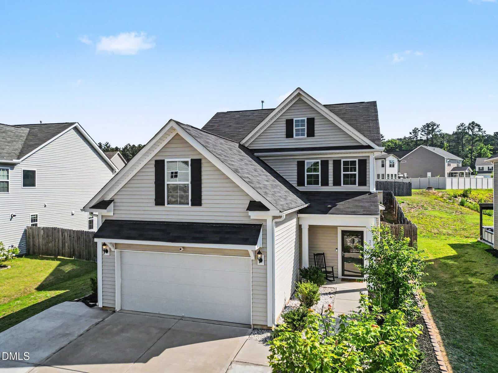 Two-story suburban house with white siding, black shutters, and attached two-car garage. Small front porch with chairs, tidy front yard, and neighboring houses visible under a blue sky.