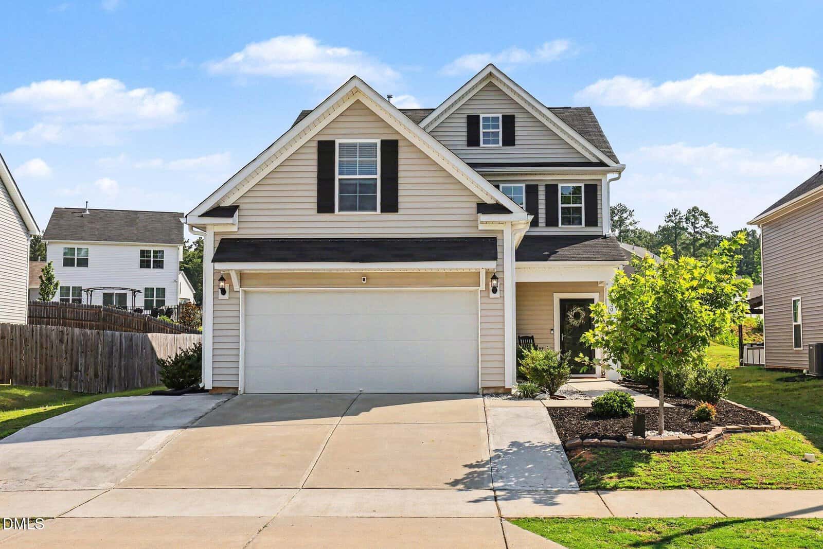 Two-story suburban house with beige siding, black shutters, and a double garage. The driveway is wide and clean, and there is landscaping with small shrubs and a tree near the front porch. The sky is clear and sunny.