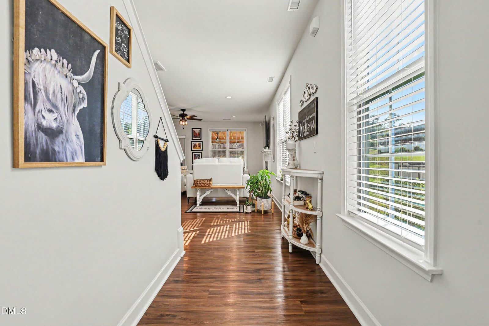 A bright hallway with wood floors, white walls, large windows with blinds, and wall art, leading to a sunlit living room with a white sofa, plants, and a ceiling fan.