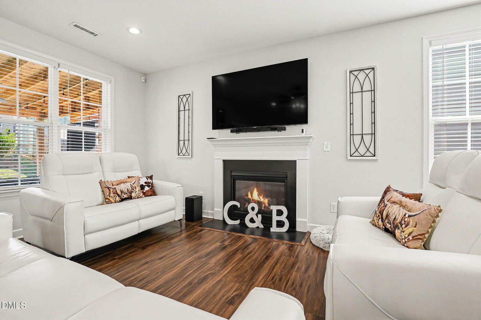 Modern living room with white sofas, a fireplace, large wall-mounted TV, decorative “C & B” letters on the hearth, and wood flooring. Two windows let in natural light.