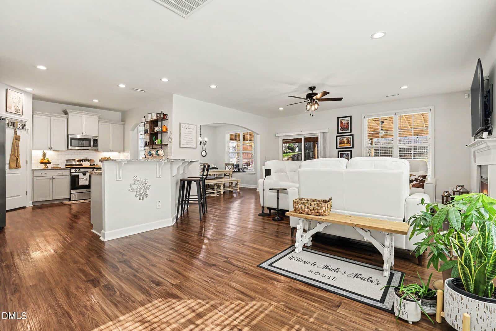 Bright open-concept living room and kitchen with white cabinetry, wood floors, white furniture, ceiling fan, wall art, indoor plants, and large windows allowing natural light. Cozy and inviting home interior.