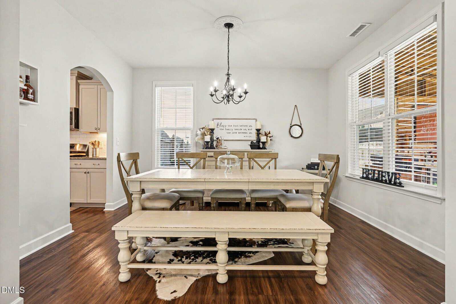 A bright dining room with a long farmhouse table, bench seating, six chairs, chandelier, wood floors, large windows with blinds, and a cowhide rug. A kitchen is visible through an arched doorway.