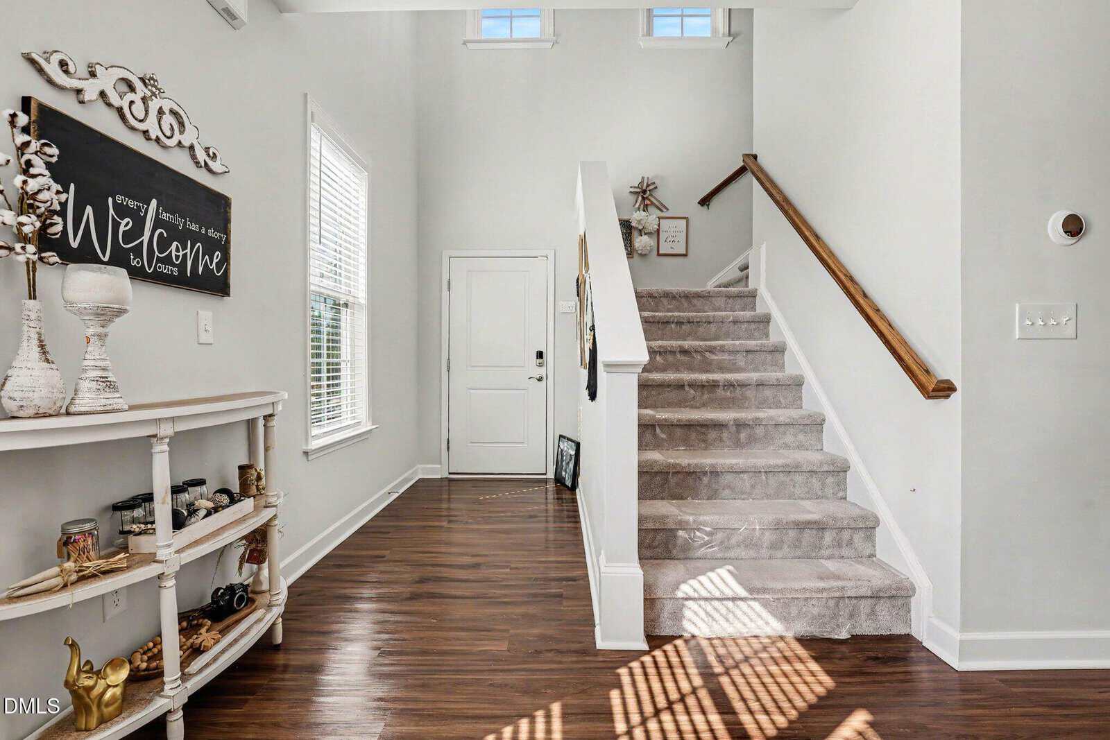 Bright entryway with dark wood floors, carpeted stairs, a white door, and a decorative console table displaying jars, books, and a welcome sign. Sunlight streams through window blinds onto the floor.