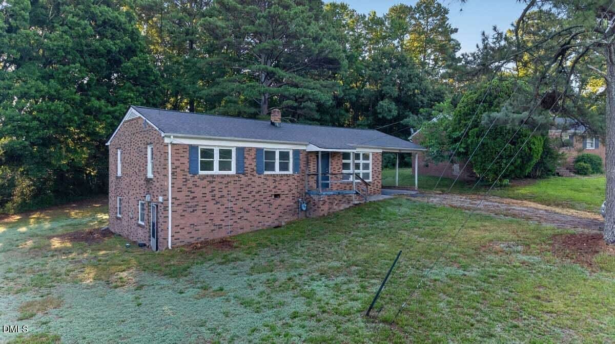 A single-story brick house with blue shutters and a front porch sits on a sloped lawn, surrounded by trees. A carport is attached on the right side of the house.