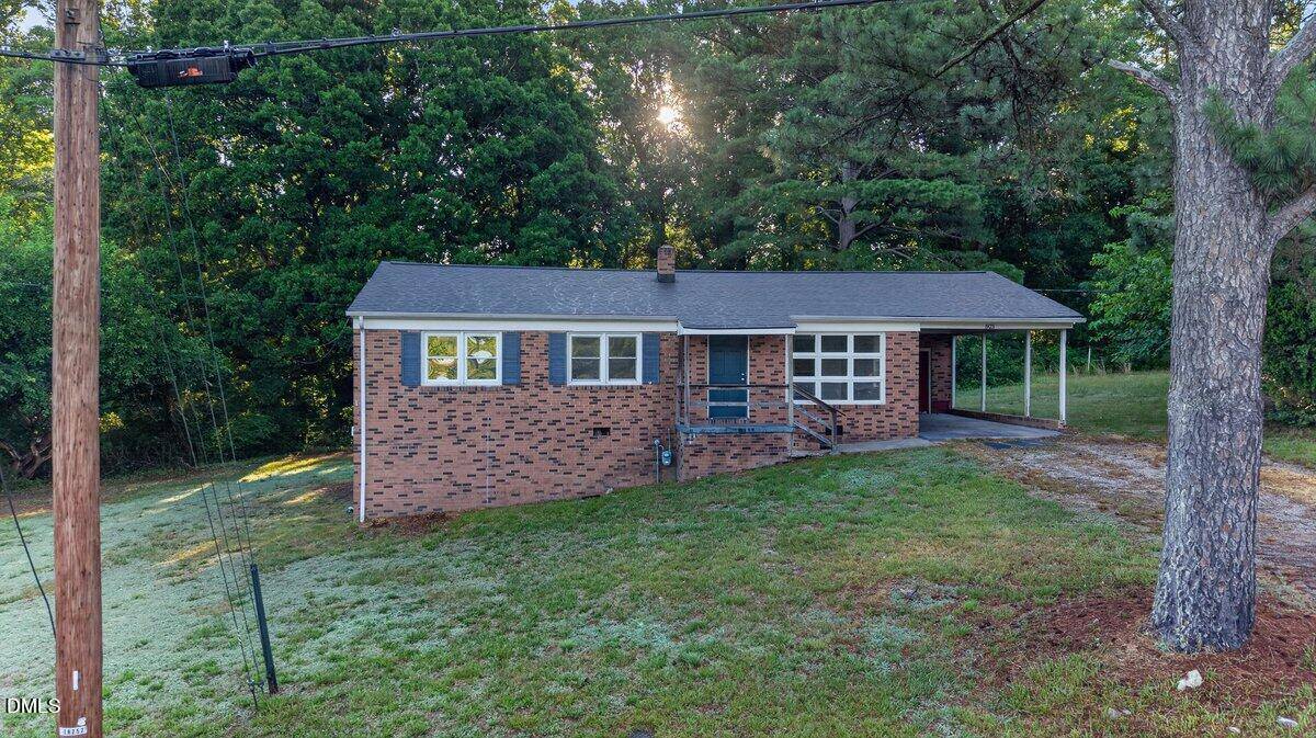 A single-story brick house at 921 Patton Circle in Henderson features a front porch, carport, and sloped yard, surrounded by tall trees and greenery. Sunlight filters through the trees in the background.