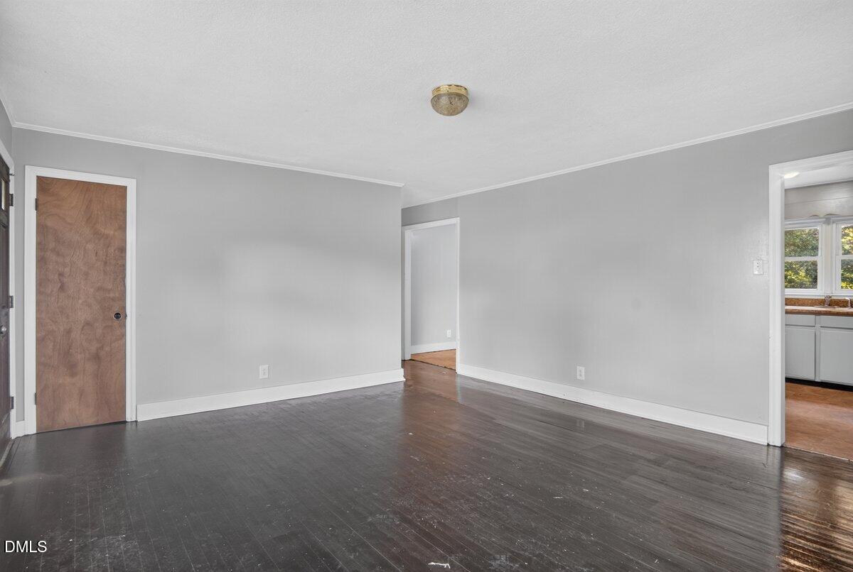 Empty room with dark wood floors, gray walls, white trim, and a ceiling light fixture. Door on the left, hallway in the center, and an opening to a bright kitchen area on the right.