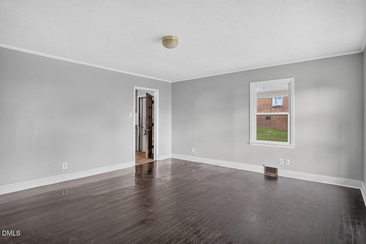 Empty room with gray walls, dark wood floors, a white-trimmed window showing a brick building and green grass outside, white ceiling with a central light fixture, and a doorway leading to another area.