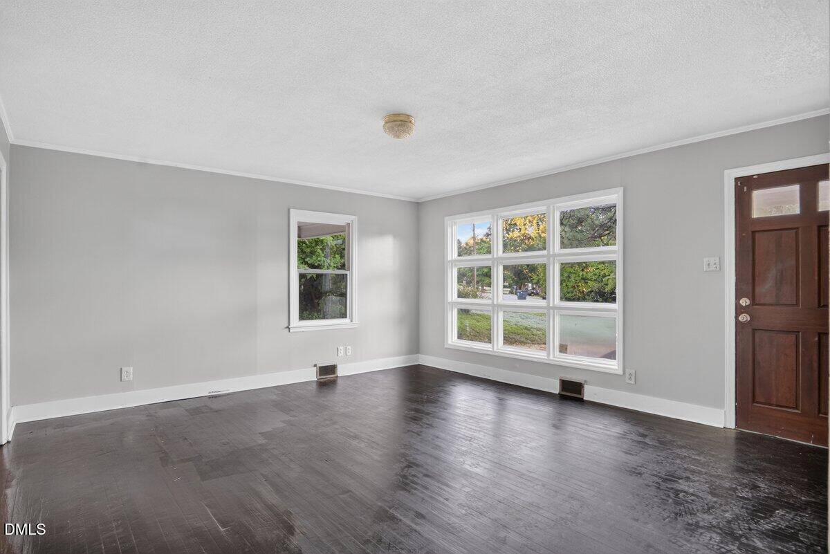 Bright, empty living room with dark hardwood floors, light gray walls, a wooden front door, large windows letting in natural light, and a ceiling light fixture.
