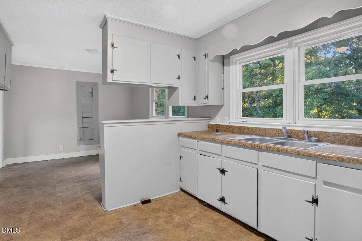 A kitchen with white cabinets, brown countertops, a double sink under two large windows, and a partial wall dividing it from an adjacent dining area.