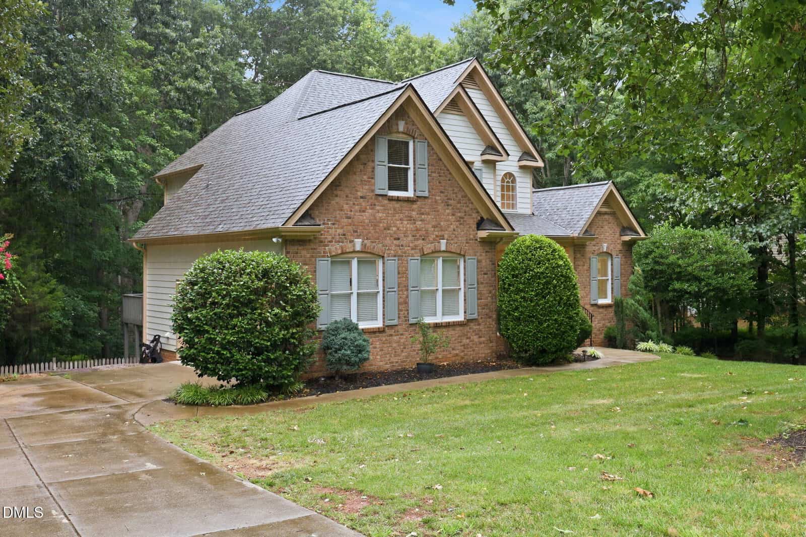 A two-story brick house with a steep gray roof, white trim, and multiple front-facing gables, surrounded by green shrubs and trees, with a curved concrete driveway in front.