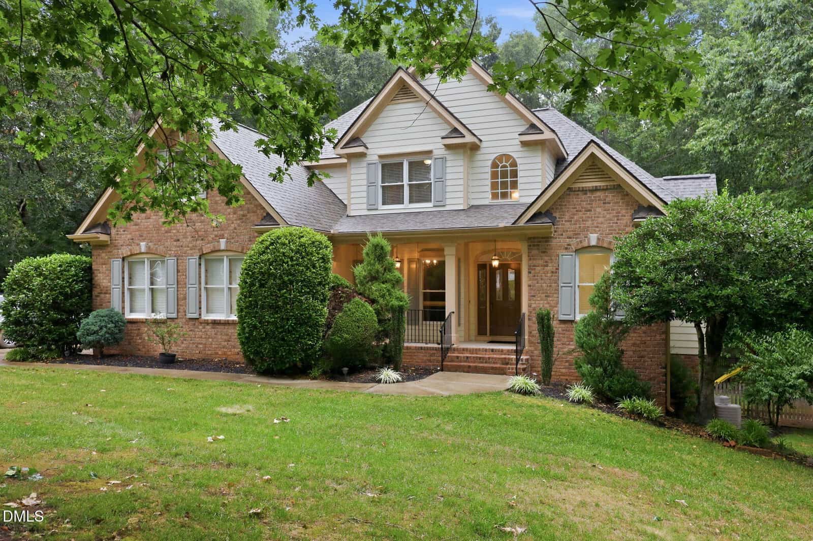 A two-story brick house with white trim, multiple front-facing gables, large windows, and a covered porch at 155 Georgetown Woods Drive in Youngsville. The home is surrounded by manicured bushes, trees, and a grassy yard.