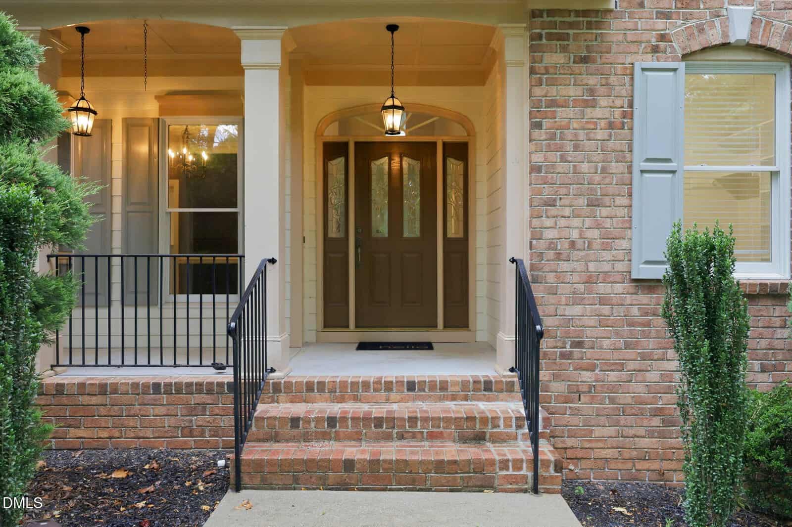 A brick house entryway with double front doors, glass panels, and a black metal railing along the steps. There are two hanging lights and a window with pale blue shutters on the right. Shrubs line the walkway.