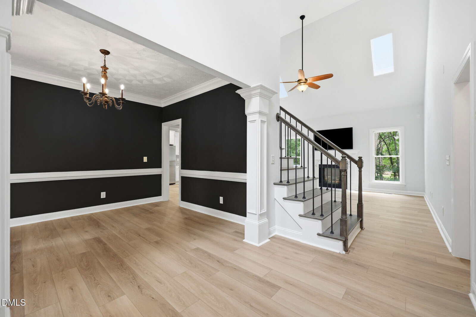 A modern home interior with light wood floors, white trim, black accent walls in the dining area, a staircase with a metal railing, a ceiling fan, large windows, and high ceilings with a skylight.