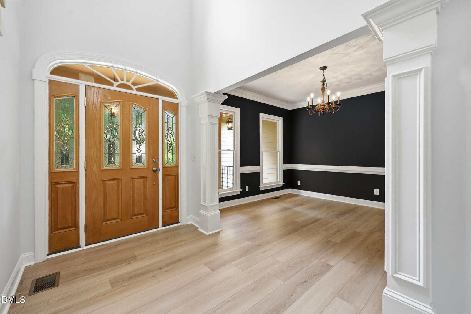 A bright entryway with double wooden doors featuring decorative glass, leading into a dining room with black walls, white trim, light wood floors, and a chandelier.