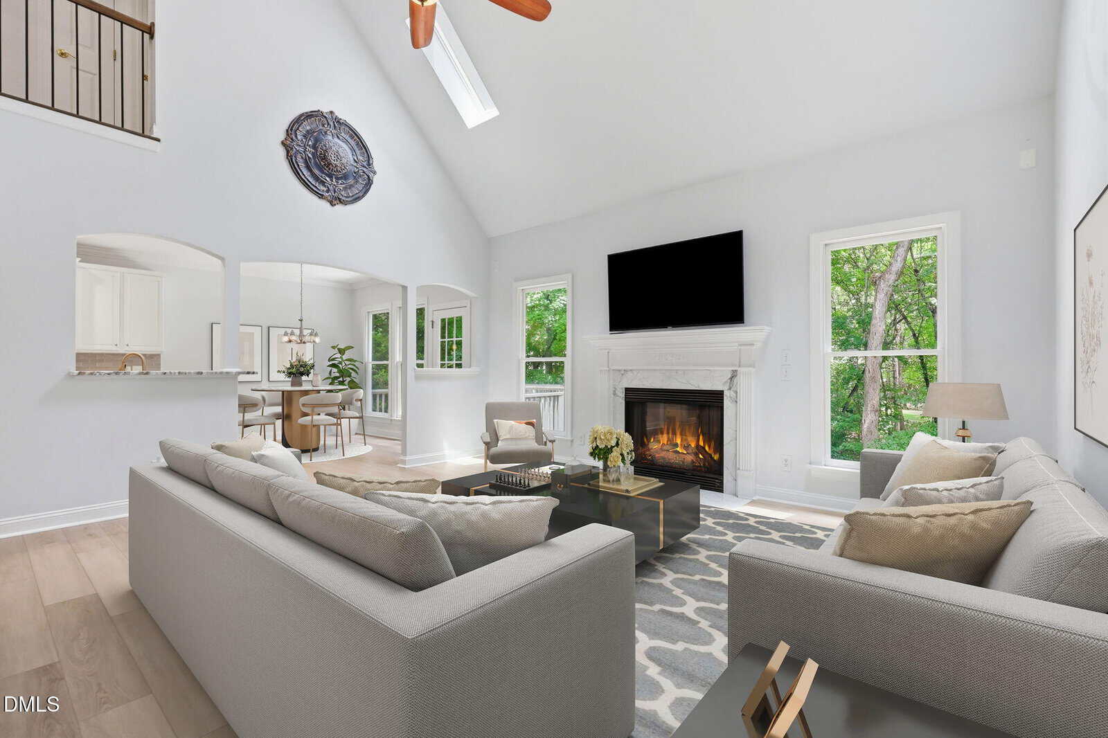 Bright living room with light gray sofas, a fireplace below a mounted TV, large windows showing greenery, and a patterned rug. Dining area and kitchen are visible through arched openings.