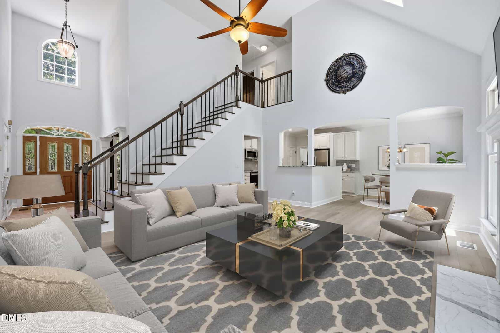 Bright living room with gray sofas, a patterned rug, a black coffee table, and a stairway leading upstairs; open kitchen and dining area visible, with large windows and vaulted ceiling.