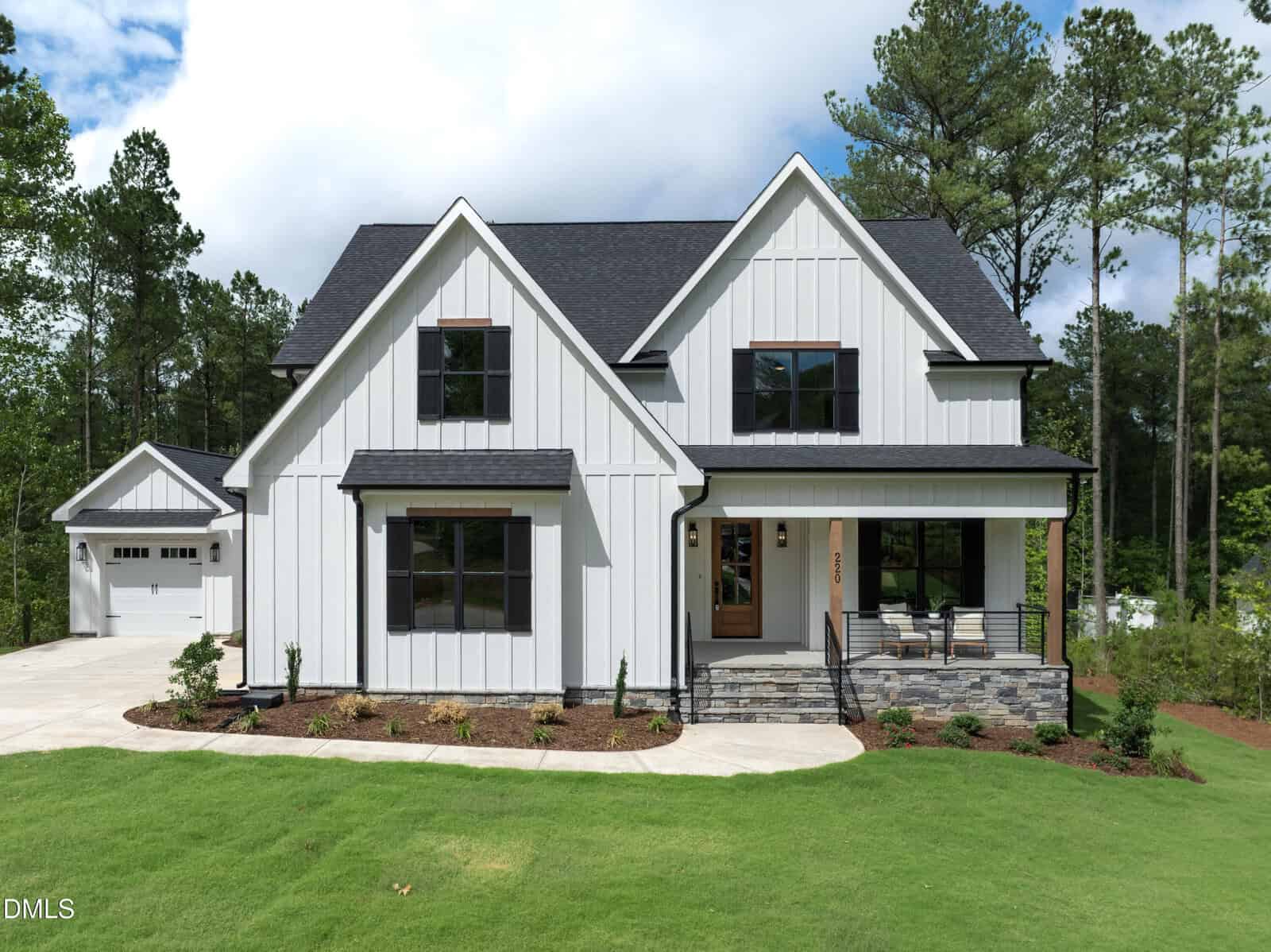 A modern farmhouse-style home at 220 Whistlers Cove in Louisburg, featuring white board-and-batten siding, black window frames, a covered front porch with stone accents, a detached garage, green lawn, and tall trees.
