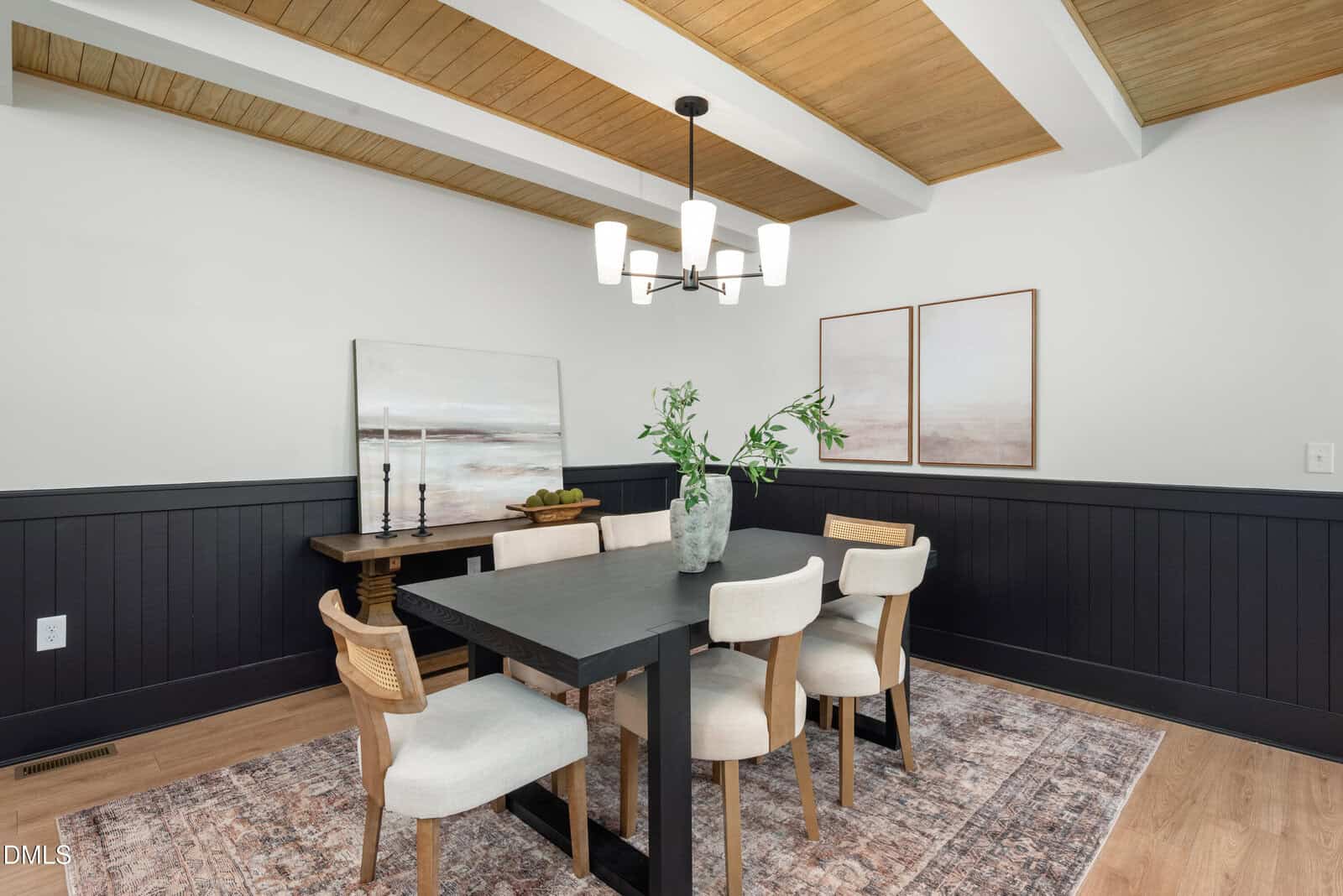 Modern dining room with a black table, six chairs, a light fixture above, decorative vases with greenery, two wall art pieces, and wood accents on the ceiling. The room features black wainscoting and a patterned rug.