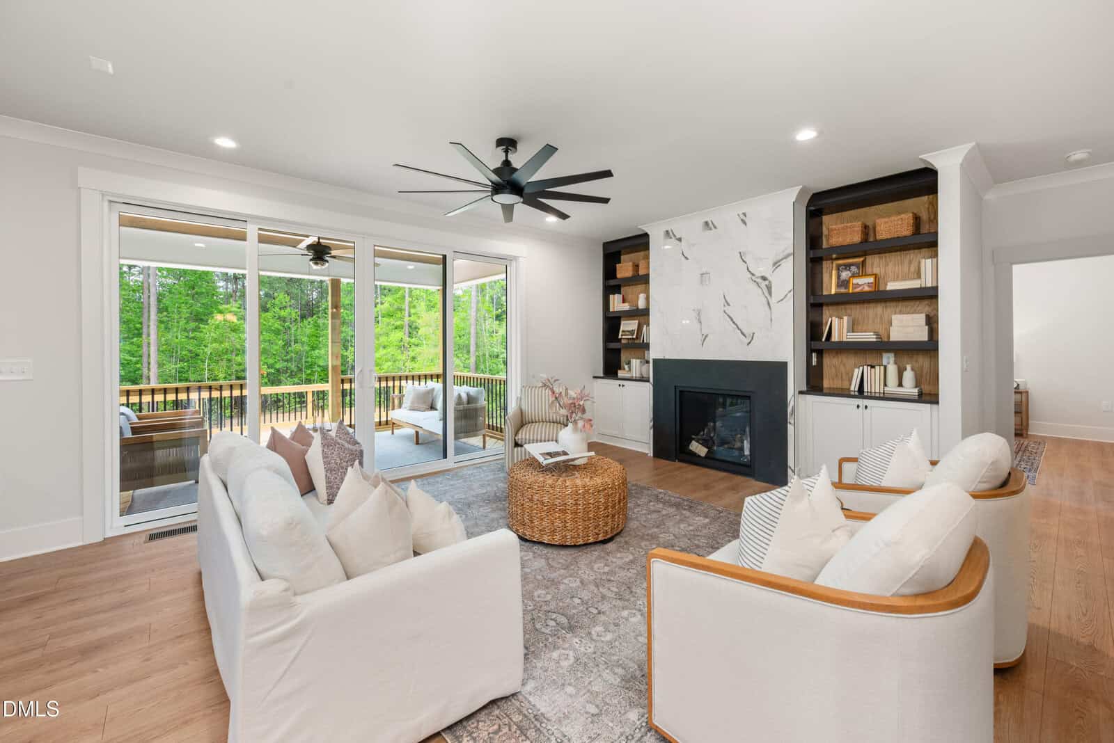 Bright living room with white walls, a modern ceiling fan, built-in shelves, a marble fireplace, two white armchairs, a white sofa, a round wicker coffee table, and large sliding glass doors opening to a deck with forest views.