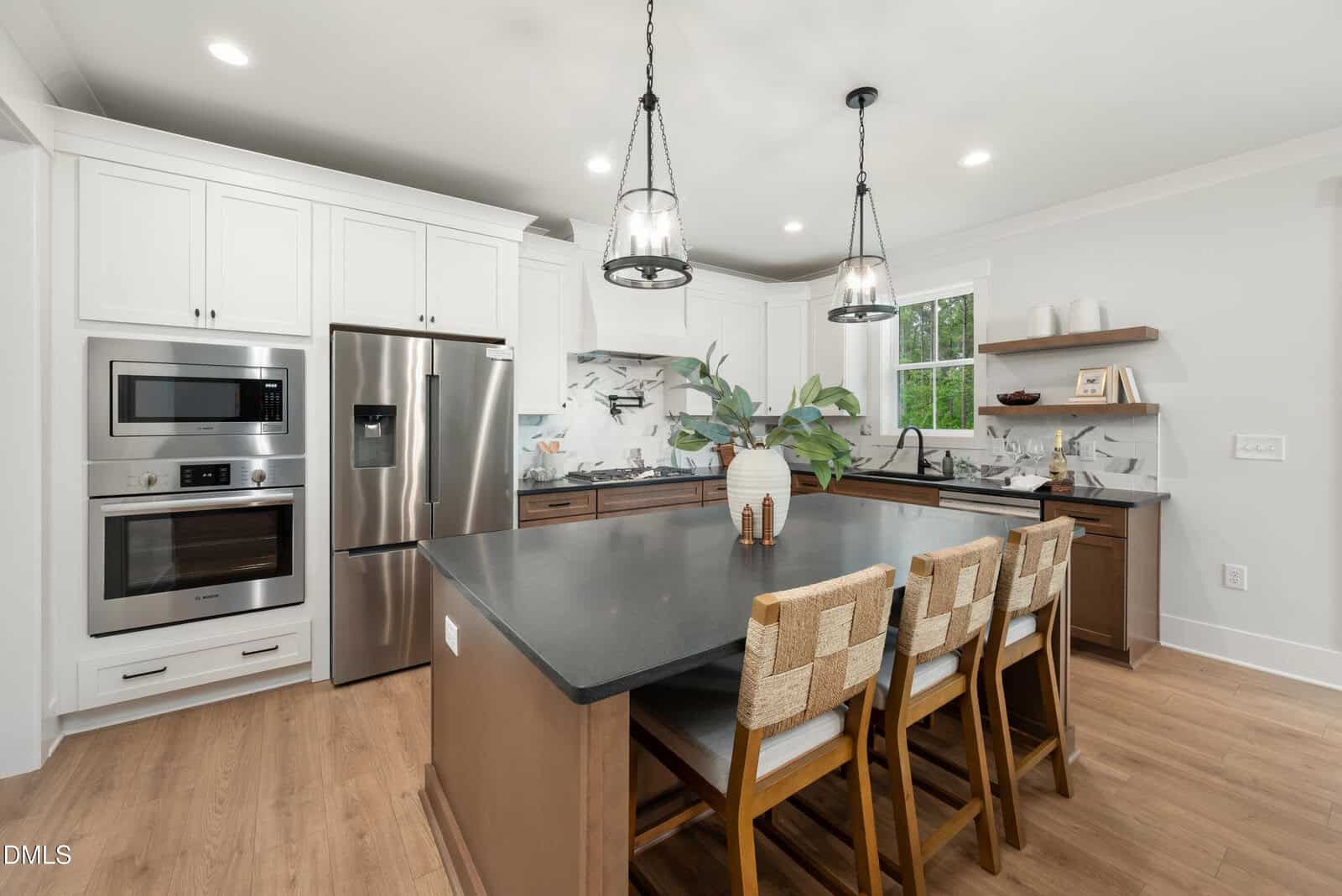 Modern kitchen with white cabinets, stainless steel appliances, a dark island with four woven-back chairs, pendant lights, wood floors, marble backsplash, open shelves, and a vase with greenery.