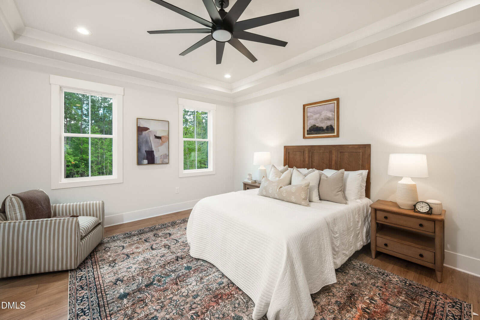 A bright bedroom with a large bed, white bedding, wooden headboard, two bedside tables with lamps, a patterned rug, two windows, wall art, and a striped armchair. Ceiling has tray details and a black ceiling fan.