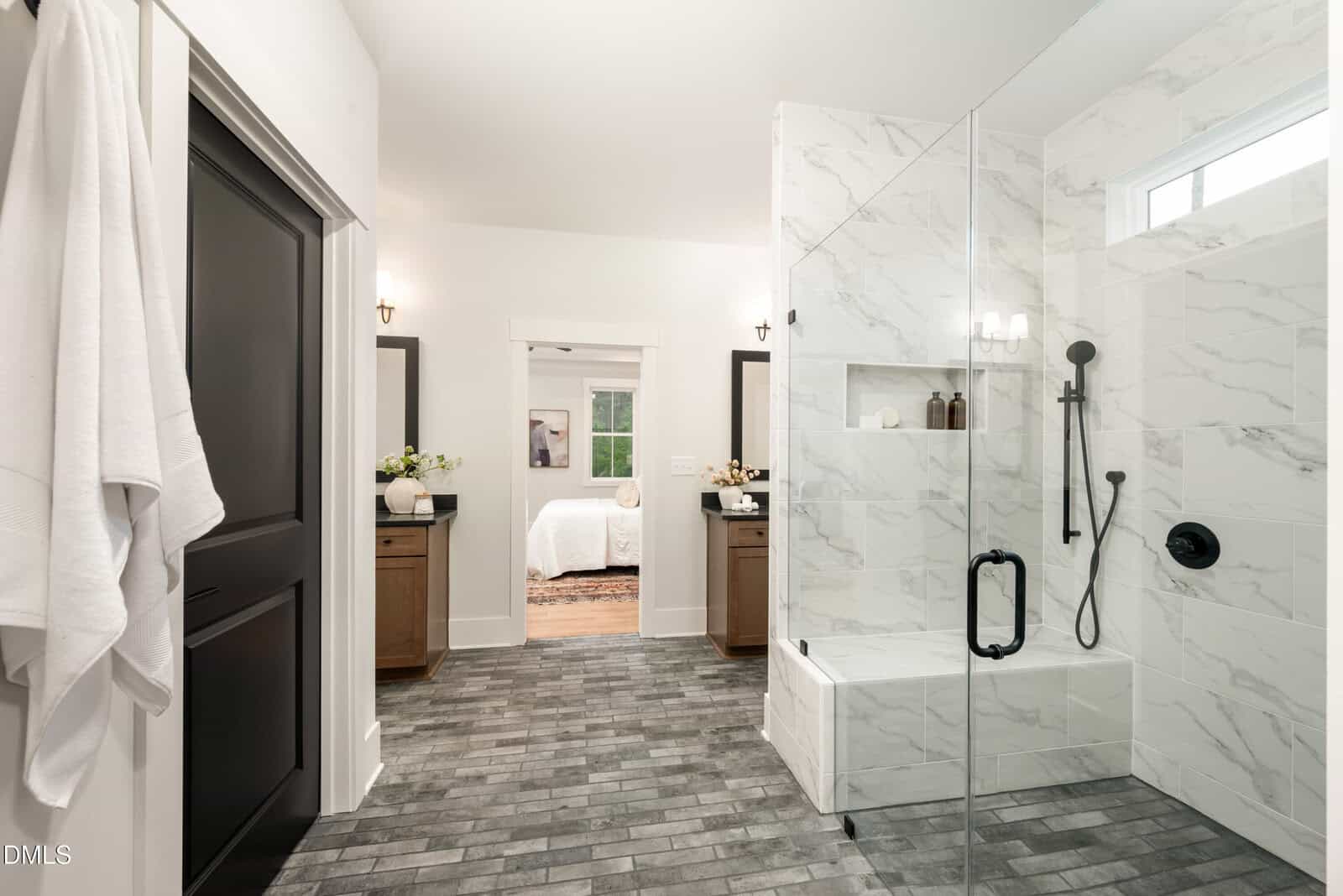 Modern bathroom with glass shower, marble walls, black fixtures, gray tile floor, double sinks with wooden cabinets, white towels hanging by a dark door, and view into a bedroom with large window.