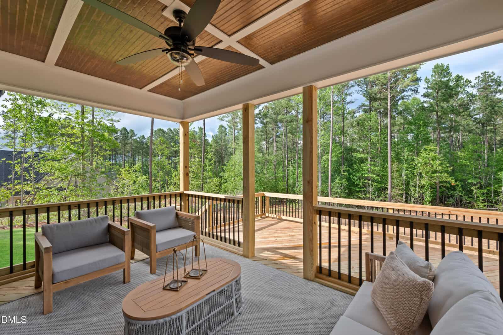 A cozy covered porch with modern patio furniture overlooks a wooden deck and lush green trees, creating a peaceful outdoor living space.