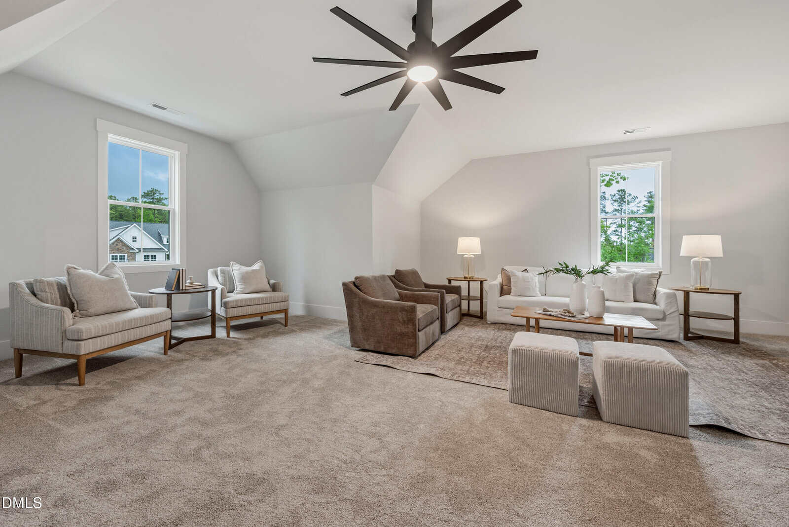 Spacious living room with neutral decor, two seating areas, modern armchairs, sofas, large windows, and a black ceiling fan. Beige carpet and white walls create a bright, airy atmosphere.