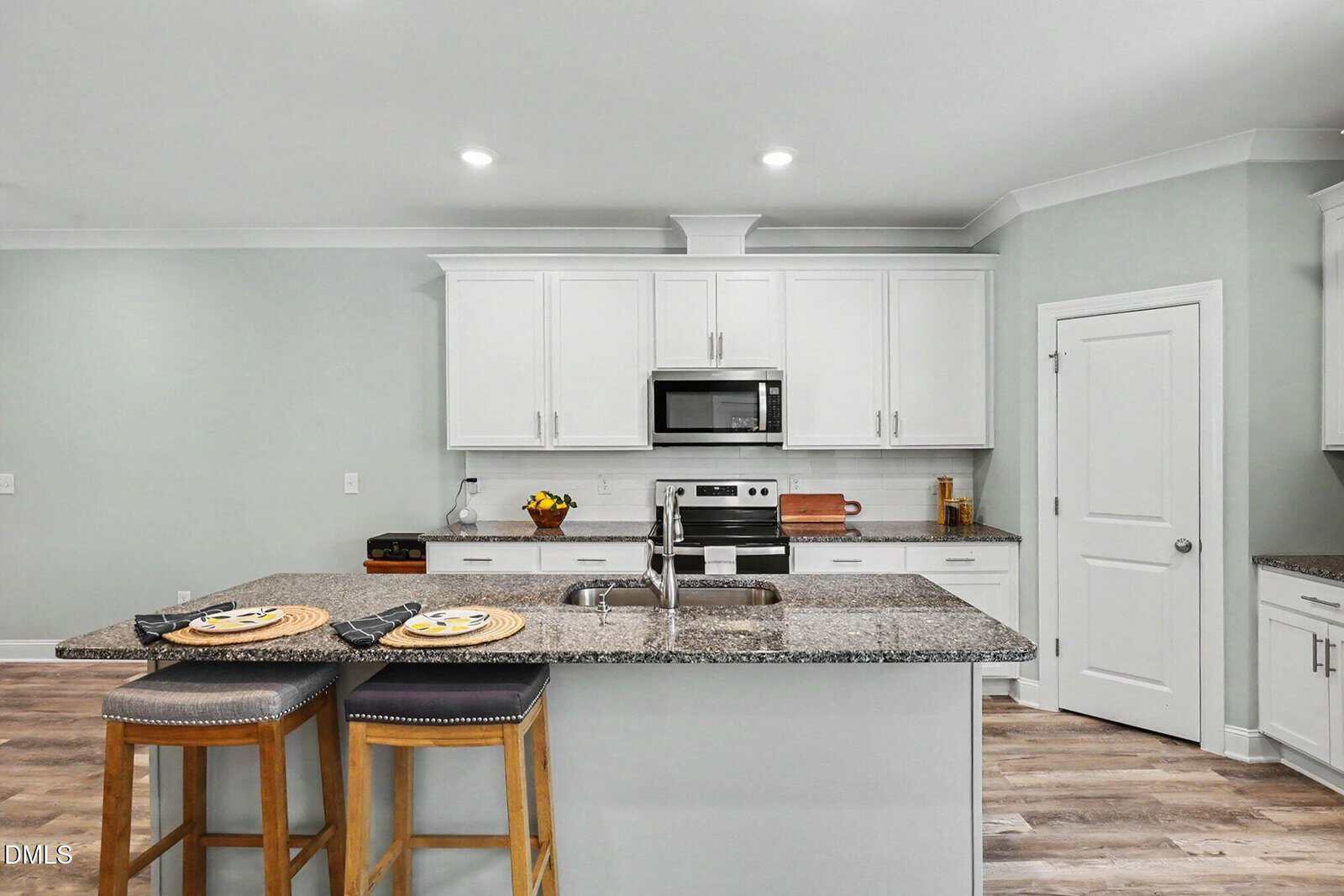 Modern kitchen with white cabinets, granite countertops, stainless steel appliances, and an island with two stools. The floor is wood, and the walls are painted light gray.