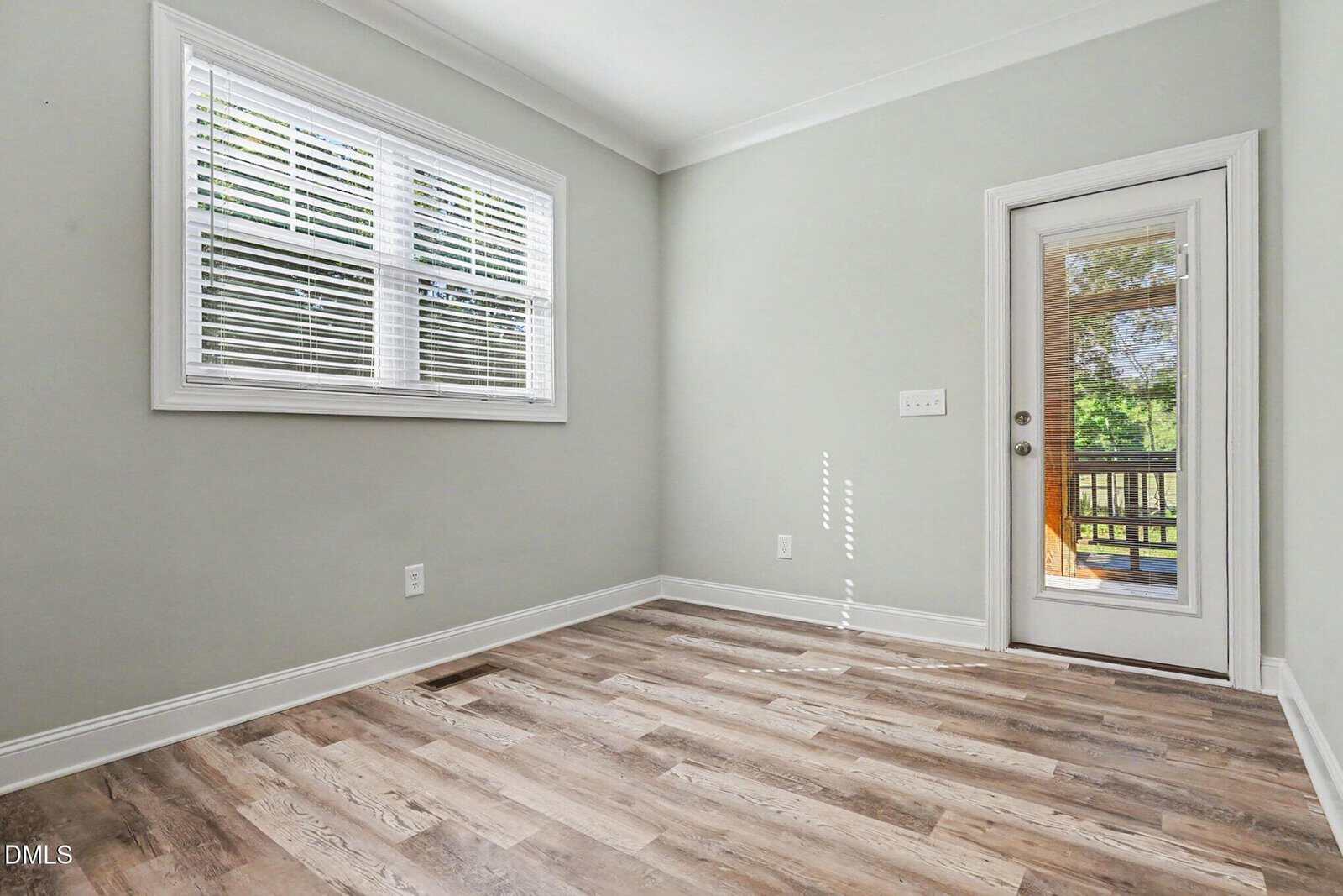 Bright, empty room with light gray walls, wood-patterned flooring, a double window with white blinds, and a glass door leading outside to a deck with green trees visible.