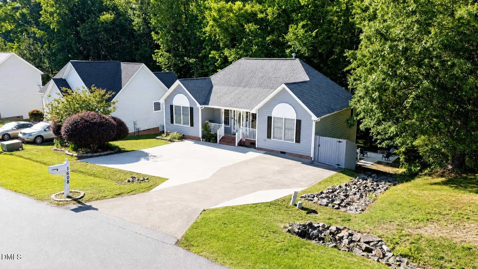 Single-story gray house with white trim, a covered front porch, and a large driveway. The yard is well-maintained with some trees and shrubbery. Neighboring homes and a mailbox are also visible.