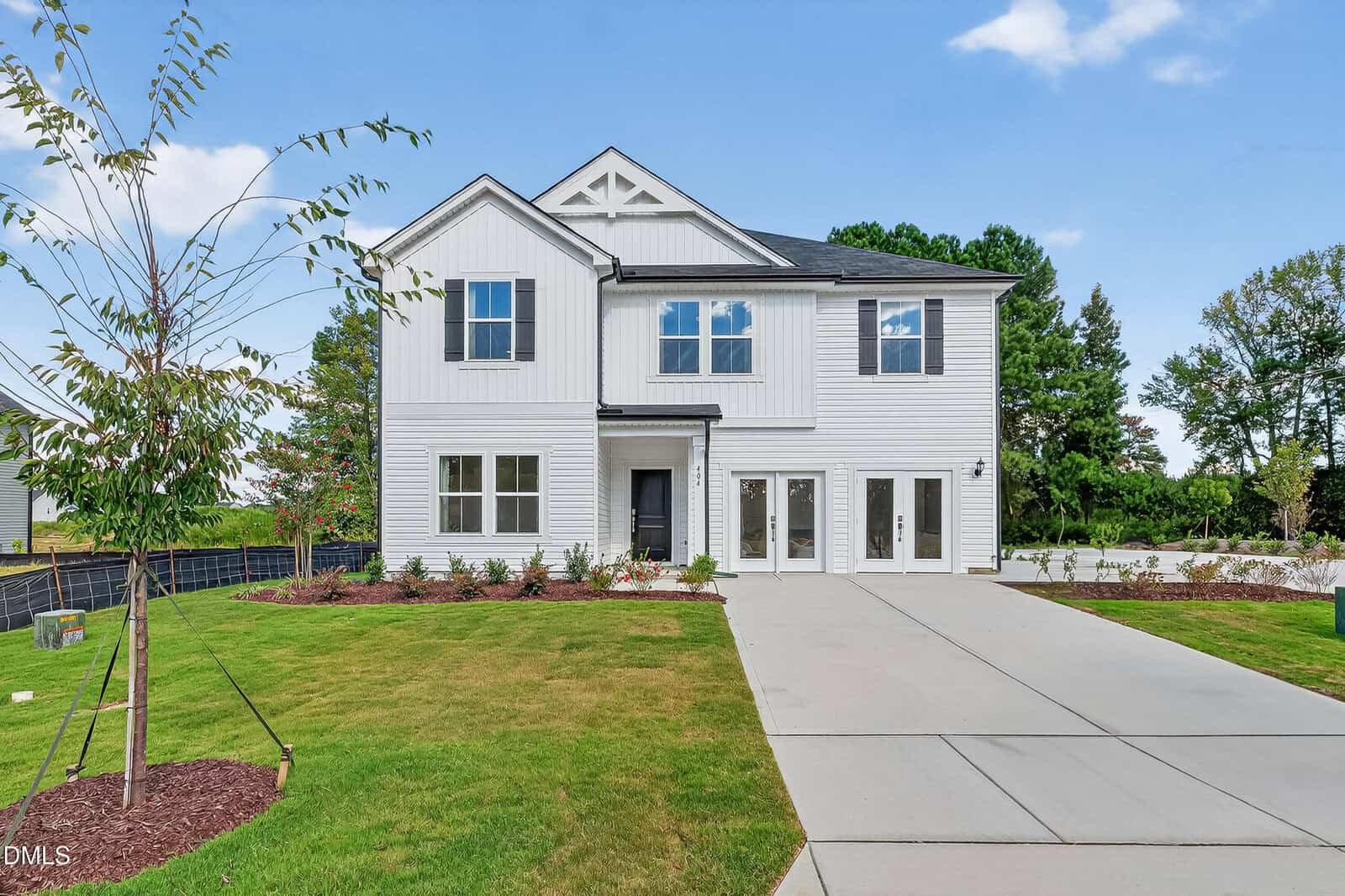 Modern two-story white house with black trim at 111 Airdale Drive, Oxford, featuring a double garage, large driveway, and landscaped front yard with young trees and shrubs against a backdrop of green trees and blue sky.