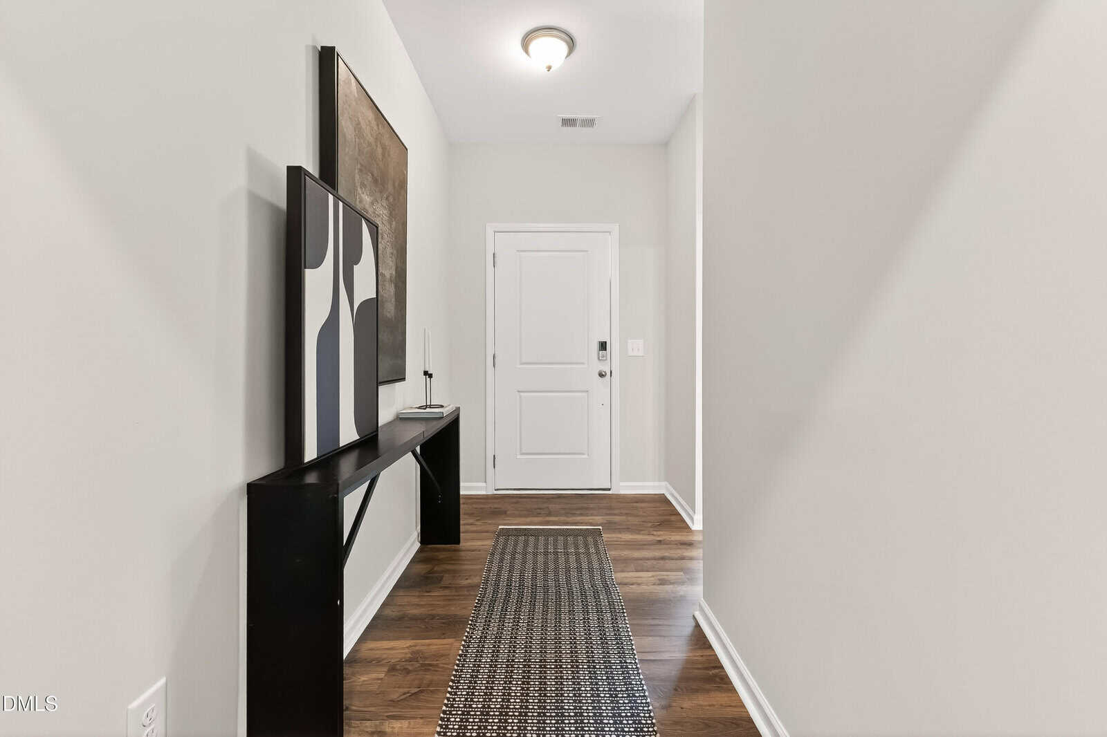 A hallway with light-colored walls, wood flooring, a white door at the end, a narrow black console table on the left with abstract art above and on it, and a textured runner rug along the floor.