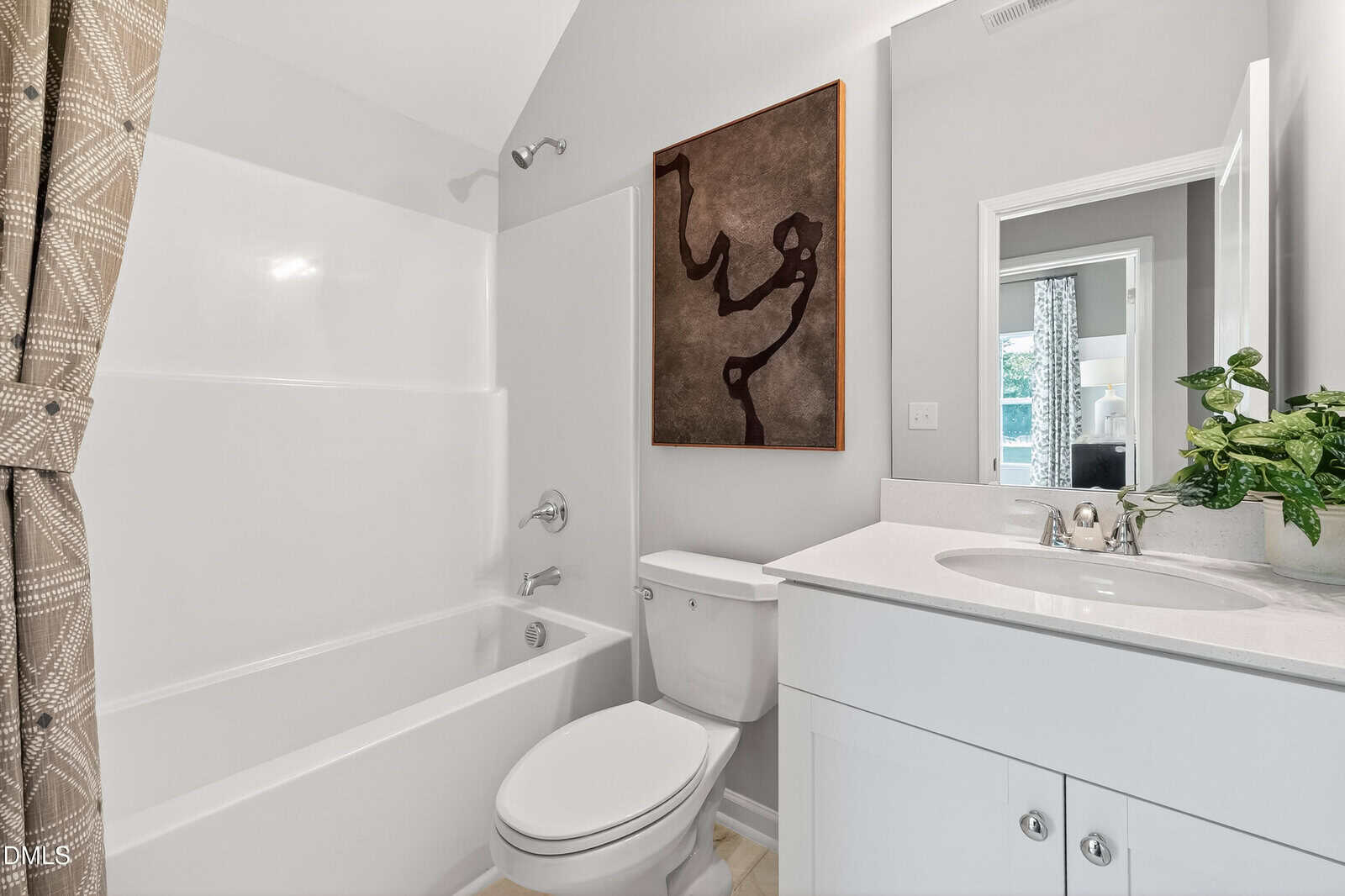 A modern bathroom with a white bathtub-shower combo, a toilet, a white vanity with a sink, a plant, and abstract wall art above the toilet. A patterned shower curtain is partly visible to the left.