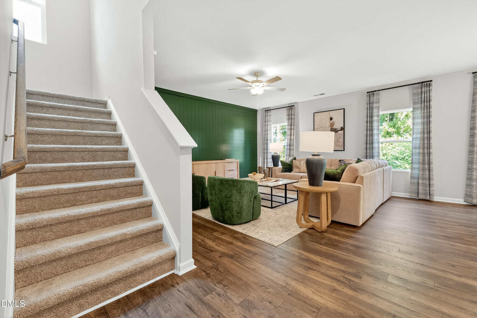 A modern living room with wood flooring, beige and green furniture, a green accent wall, large windows with patterned curtains, and a carpeted staircase leading to the upper floor.