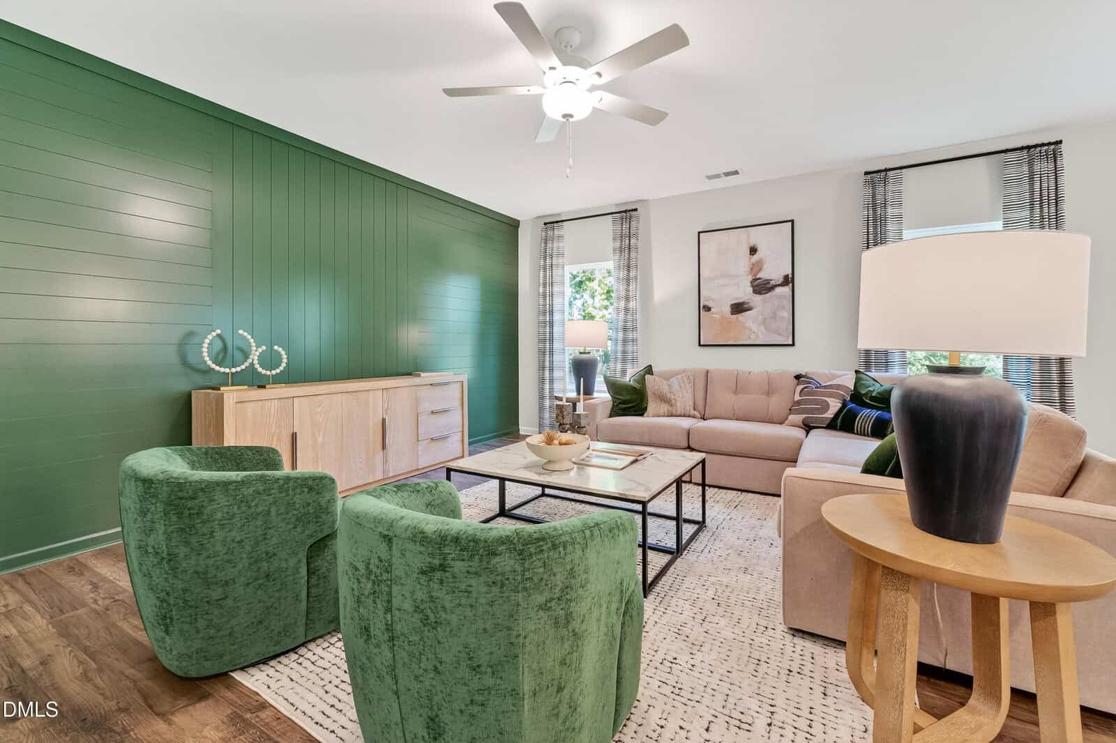 Modern living room with a green accent wall, beige sectional sofa, two green velvet chairs, wooden sideboard, abstract art, coffee table, and floor lamp, with neutral decor and natural light from windows.