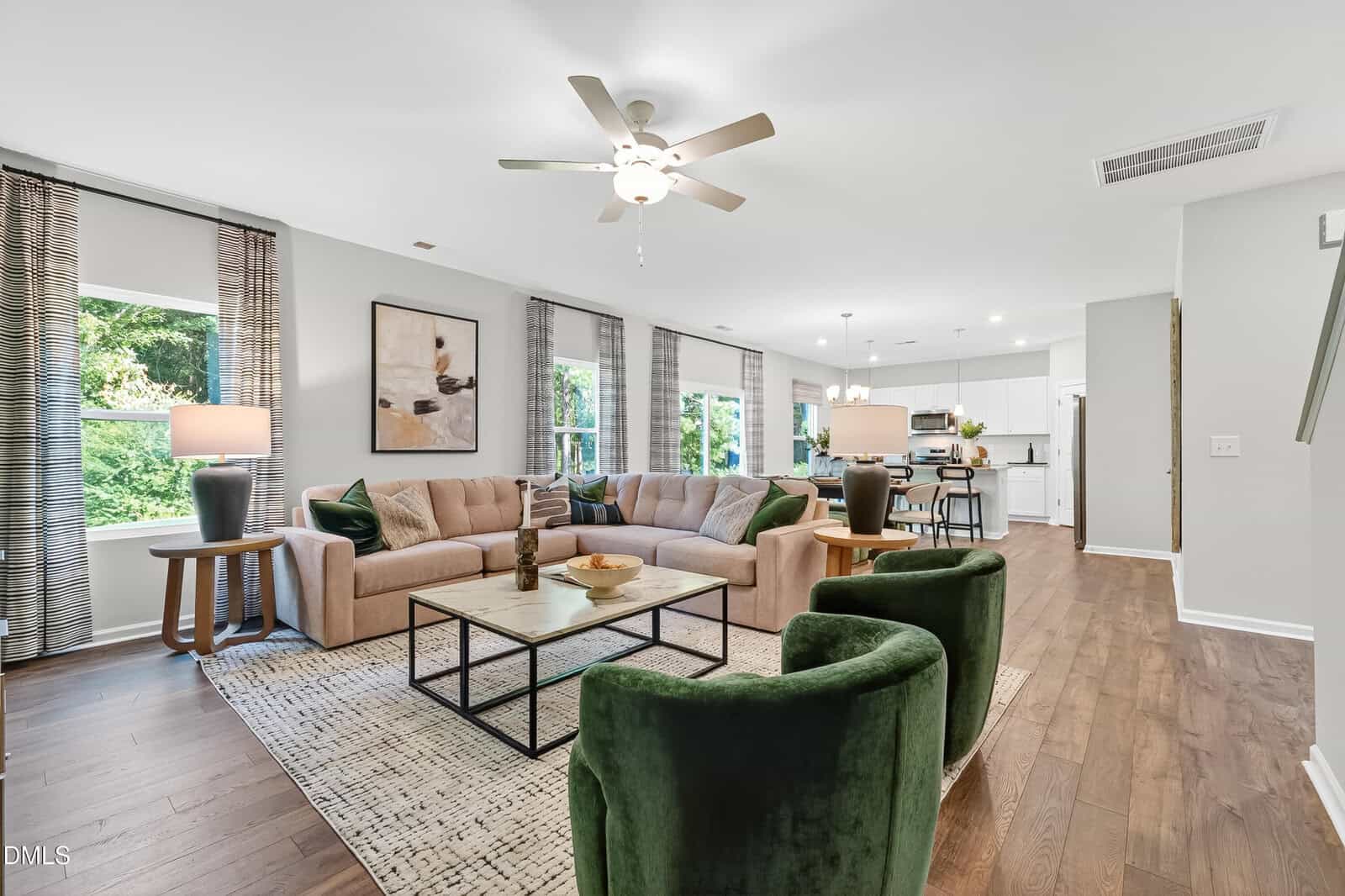 Modern living room with a tan sectional sofa, two green velvet chairs, wood floors, large windows with drapes, and a view into a white kitchen and dining area. Ceiling fan and stylish decor complete the space.