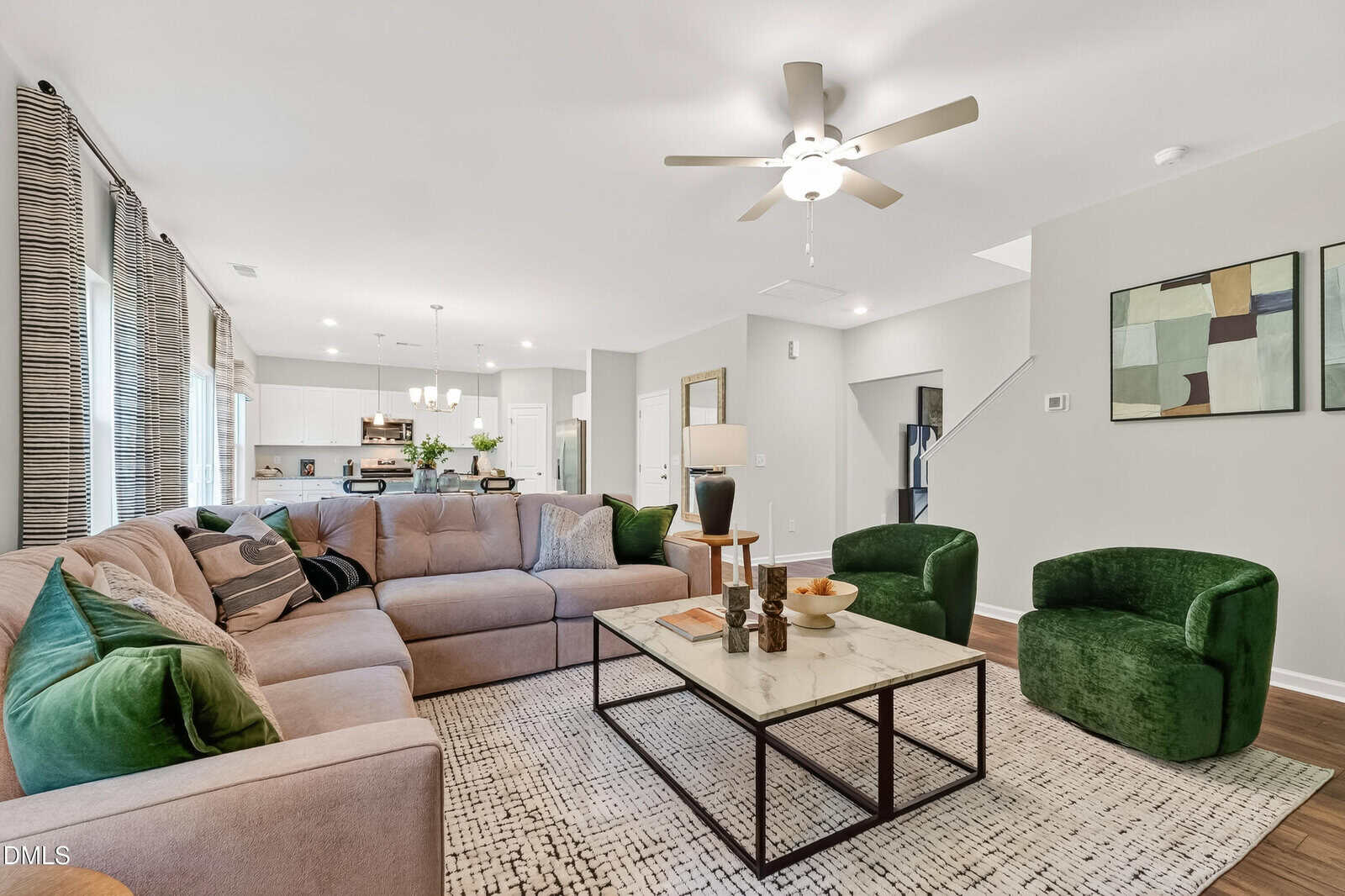 Modern living room with a large sectional sofa, two green velvet chairs, a marble coffee table, and wall art. The space is bright with neutral walls and an open view into the kitchen and dining area.