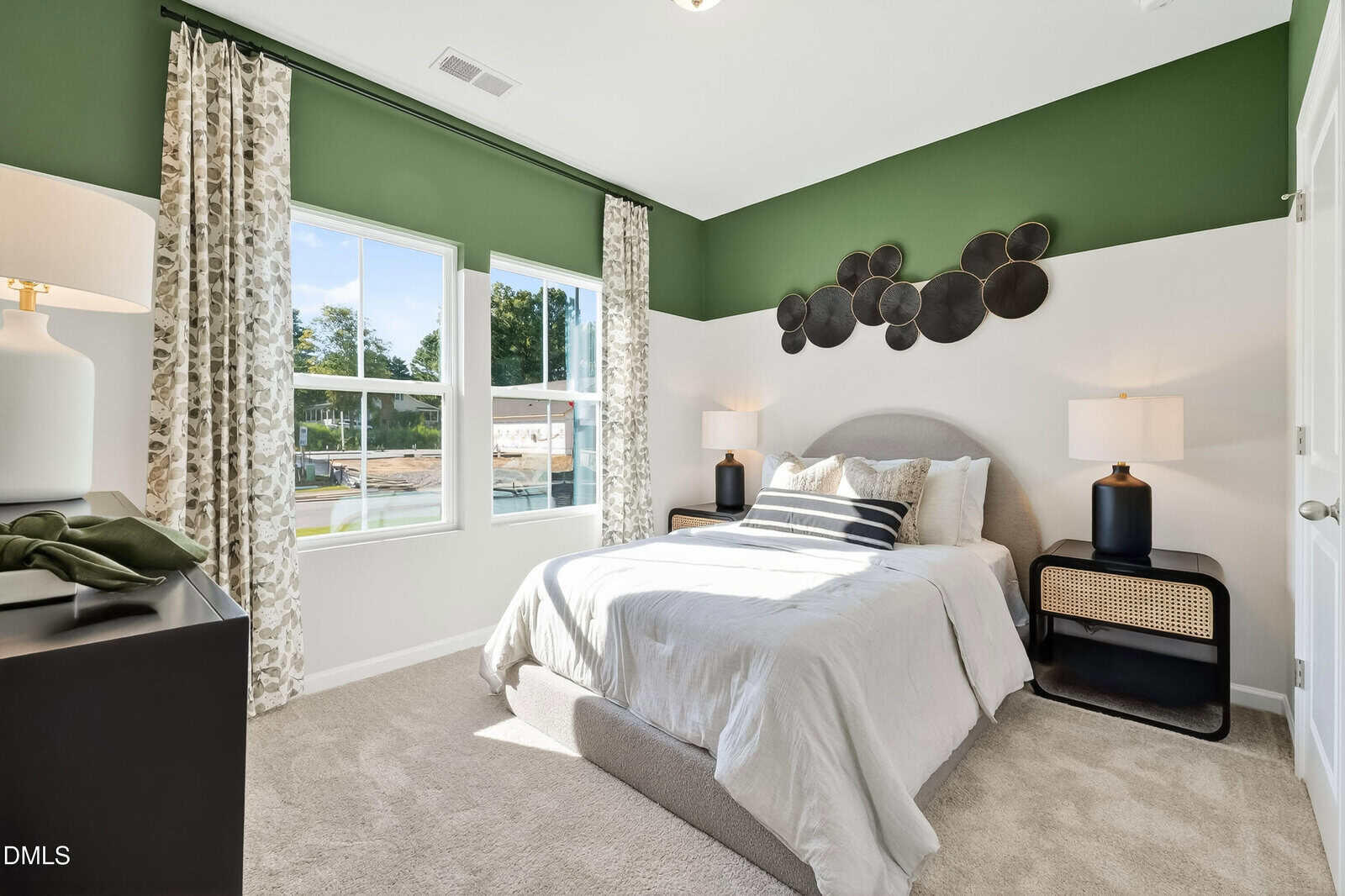A bright bedroom with green and white walls, large windows with floral curtains, a grey upholstered bed, two black lamps on matching nightstands, and circular black wall art above the headboard.
