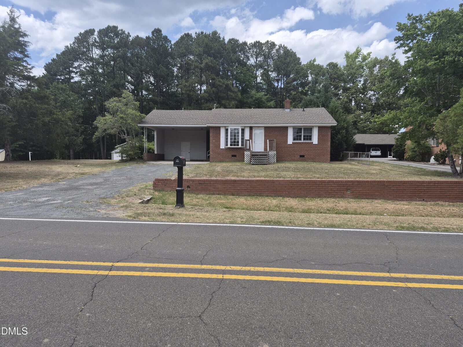 A small brick house with white trim, a porch, and an attached carport sits behind a low brick wall along a two-lane road. Large trees surround the property, and the yard is mostly grass.